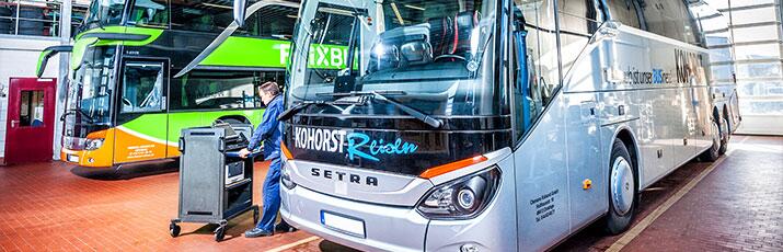 Bus-Mechatroniker mit Werkstattwagen arbeitet an einem Bus, zweiter Bus im Hintergrund.