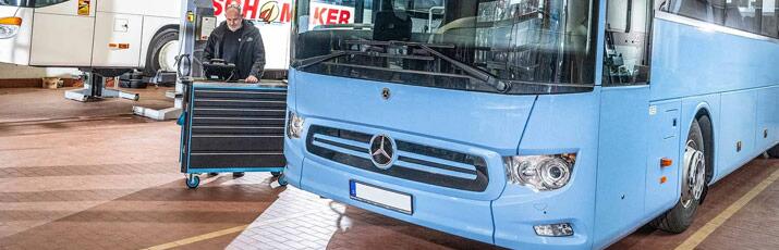Blauer Mercedes-Bus in der Werkstatt, dahinter Mechatroniker mit Werkzeugwagen.