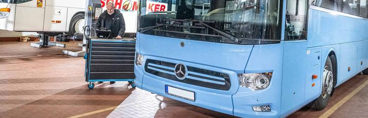 Blauer Mercedes-Bus in der Werkstatt, dahinter Mechatroniker mit Werkzeugwagen.