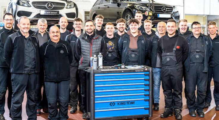 Werkstattteam in Kfz-Werkstatt vor Fahrzeugen und einem blauen Werkzeugwagen.