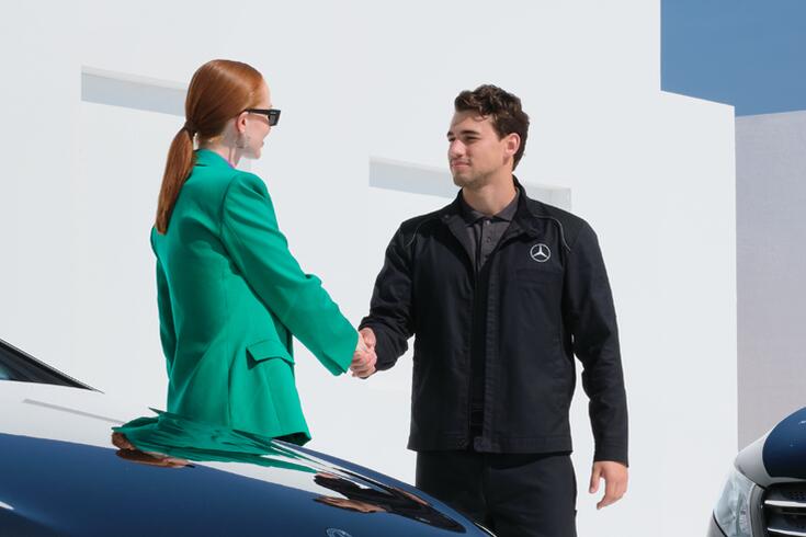 Mercedes-Benz Serviceberater gibt Kundin die Hand, im Vordergrund Front einer Limousine.